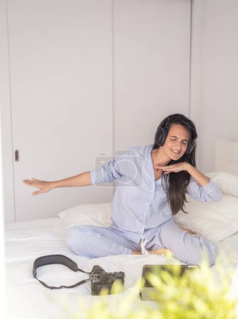 Téléchargez les photos : Femme dansante positive fermant les yeux et écoutant de la musique dans les écouteurs assis sur le lit avec tablette et appareil photo - en image libre de droit