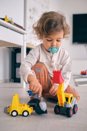 Téléchargez les photos : Curieux petit garçon avec sucette bleue jouant avec chargeur de jouets en plastique tout en étant assis sur le sol dans le salon à la maison - en image libre de droit