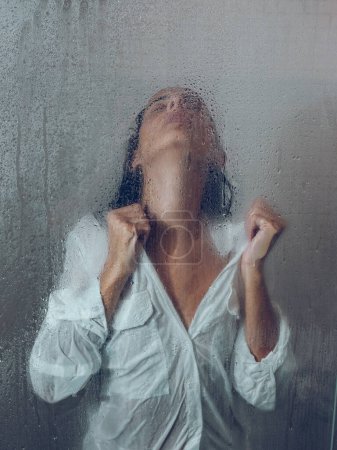 Téléchargez les photos : Grâce à un verre de femme attrayante en chemisier humide debout dans la cabine de douche légère avec porte transparente et gouttes d'eau dans la salle de bain - en image libre de droit