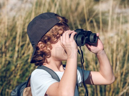 Photo for Blond boy with cap and backpack admiring nature through binoculars while on blurred background of tall grass near Pollensa town on sunny day in Mallorca, Spain - Royalty Free Image