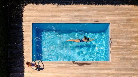 Foto de Drone vista de niño anónimo de pie en la plataforma de madera cerca de la madre joven nadando en la piscina en el día soleado durante las vacaciones de verano - Imagen libre de derechos