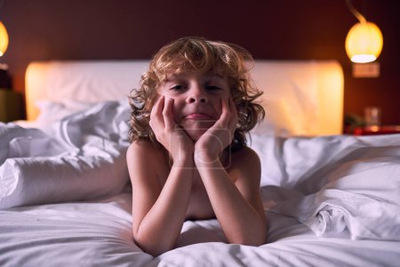 Téléchargez les photos : Mignon petit enfant aux cheveux blonds bouclés s'appuyant sur les mains et faisant visage tout en étant couché sur un lit confortable la nuit à l'hôtel - en image libre de droit