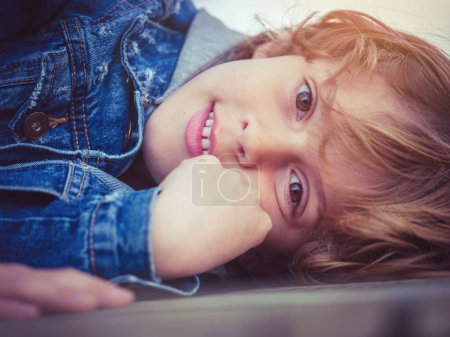 Foto de Adorable niño en chaqueta de mezclilla con cabello rubio y ojos marrones tocando la cara y mirando a la cámara mientras está acostado en el suelo - Imagen libre de derechos