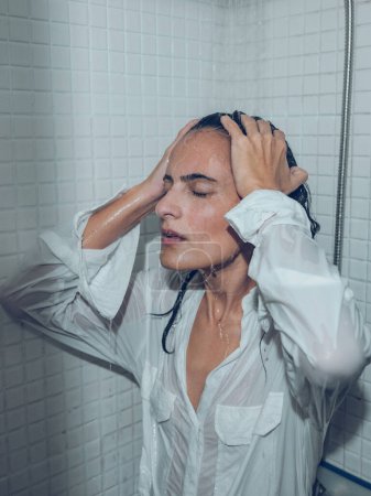 Foto de Mujer seria en camisa blanca tocando el cabello mojado mientras toma la ducha en cabina y relajante - Imagen libre de derechos