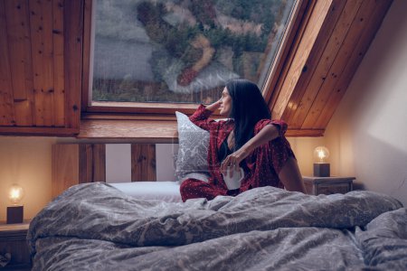 Photo for Peaceful female in pajama sitting on soft bed in cozy bedroom in house and admiring mountainous forest out of window - Royalty Free Image