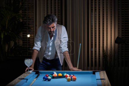 Téléchargez les photos : Homme sérieux en vêtements décontractés debout près de la table de billard avec cocktail et queue et penser au jeu tout en jouant au billard à la maison - en image libre de droit