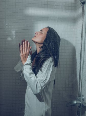 Téléchargez les photos : Vue latérale de la jeune femme avec les cheveux mouillés et chemise debout avec les yeux fermés et relaxant et méditant sous les éclaboussures d'eau de la douche peut dans la cabine - en image libre de droit