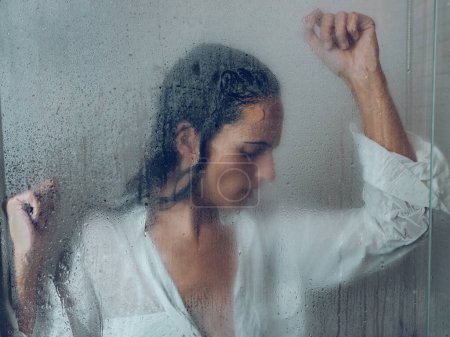 Téléchargez les photos : À travers le verre de femme désagréable avec les cheveux mouillés debout près de la porte transparente de la cabine de douche avec des gouttes d'eau dans la salle de bain légère - en image libre de droit