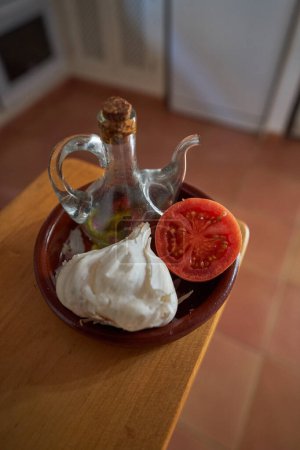 Photo for From above head of garlic with half of tomato on plate with bottle of oil in light kitchen during cooking process - Royalty Free Image