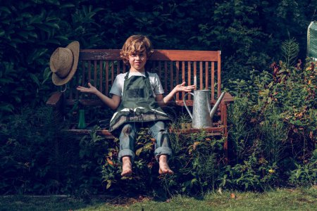 Foto de Niño triste en delantal sentado en un banco de madera con sombrero y regadera cerca de las plantas mientras mira a la cámara en verano - Imagen libre de derechos