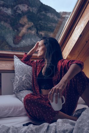 Téléchargez les photos : Femme pensive en pyjama confortable assis près de la fenêtre dans le chalet et bénéficiant d'une vue sur la forêt dans les hauts plateaux - en image libre de droit