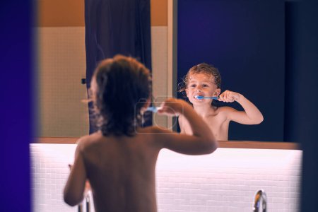 Foto de Vista posterior del lindo niño con el pelo rizado húmedo reflejándose en el espejo y cepillándose los dientes para proteger la cavidad oral - Imagen libre de derechos