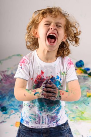 Photo for Carefree sloppy boy with dirty hands and white t shirt screaming with closed eyes while standing in messy light studio - Royalty Free Image
