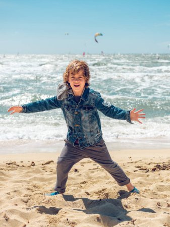 Téléchargez les photos : Joyeux petit garçon aux cheveux bouclés portant une veste en denim debout sur le sable sur le bord de la mer près de la mer ondulée avec les bras tendus et regardant la caméra - en image libre de droit