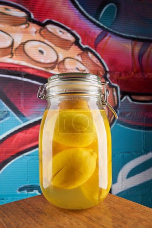 Photo for Closed glass jar with heap of ripe yellow lemons in water placed on wooden table near wall with colorful graffiti - Royalty Free Image