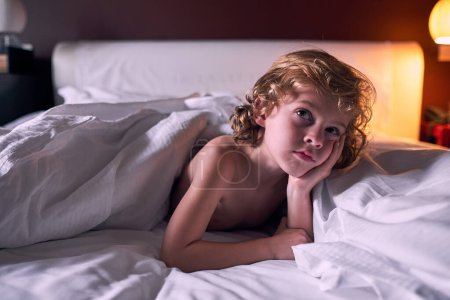 Pensive boy with curly blond hair leaning on hand and thinking while lying under warm blanket on bed in evening in hotel