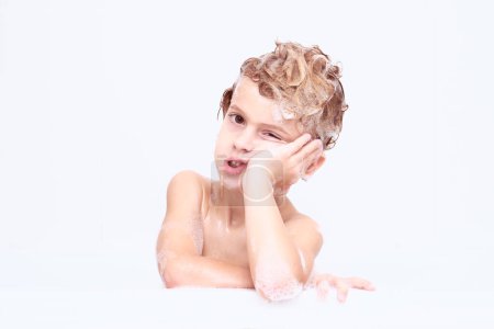 Photo for Dreary boy with soapy foam on head leaning on hand and looking at camera while washing in bathroom isolate on white background - Royalty Free Image