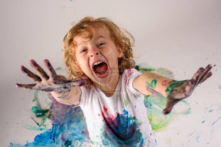 Foto de Desde arriba de chico divertido cubierto de pinturas de colores mirando a la cámara con la cara excitada en el fondo blanco en el estudio desordenado - Imagen libre de derechos