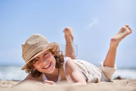 Téléchargez les photos : Joyeux préadolescent en chapeau de paille couché sur la plage et regardant la caméra tout en profitant des vacances d'été au bord de la mer - en image libre de droit