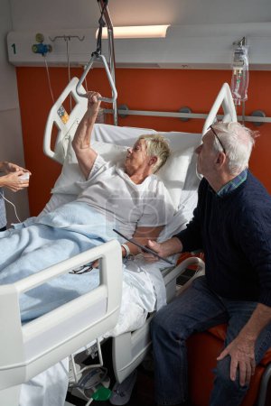 Téléchargez les photos : Patiente âgée malade avec les cheveux courts en robe médicale couchée sur le lit près du mari et communiquant avec le médecin anonyme de la culture pendant le traitement à l'hôpital - en image libre de droit