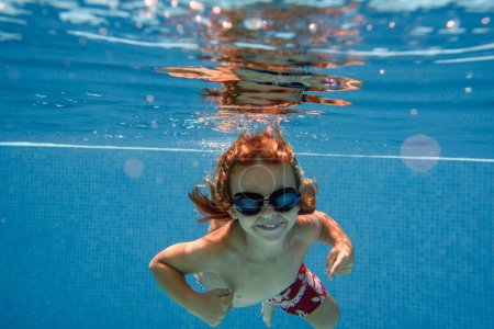 Foto de Inyección submarina de niño alegre en pantalones cortos de natación y gafas de buceo en agua limpia y transparente de la piscina y mirando a la cámara contra el fondo azul - Imagen libre de derechos