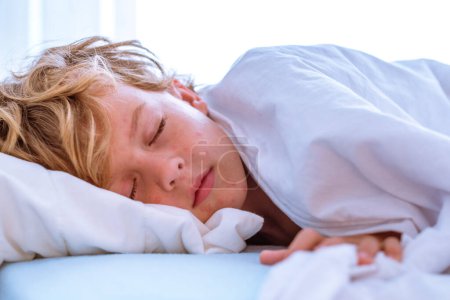 Téléchargez les photos : Garçon malade aux cheveux blonds bouclés couché sur le lit sous une couverture blanche et dormant alors qu'il souffrait de varicelle à la maison - en image libre de droit