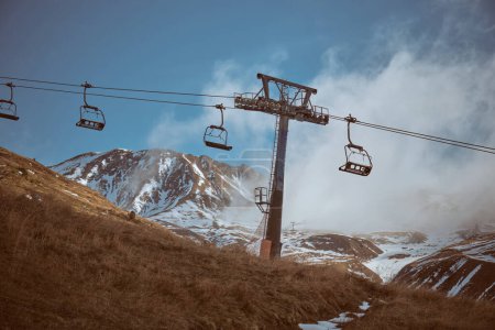 Foto de Teleférico con asientos para el transporte de turistas que van bajo colinas cubiertas de hierba rodeadas de montañas nevadas cubiertas de vapor - Imagen libre de derechos