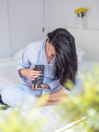 Téléchargez les photos : Femme focalisée en pyjama ajustant l'appareil photo analogique à l'ancienne assis avec les jambes croisées sur le lit le matin - en image libre de droit