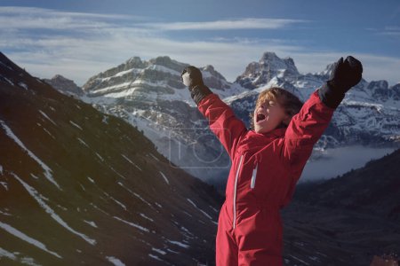 Photo for Boy in warm clothes raising arms with snowball and yelling loudly while standing against snowy mountain ridge on sunny day - Royalty Free Image