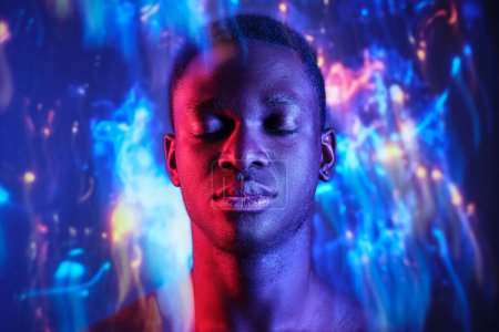 Téléchargez les photos : Calme homme afro-américain aux épaules nues debout avec les yeux fermés dans des lumières floues en studio sombre - en image libre de droit
