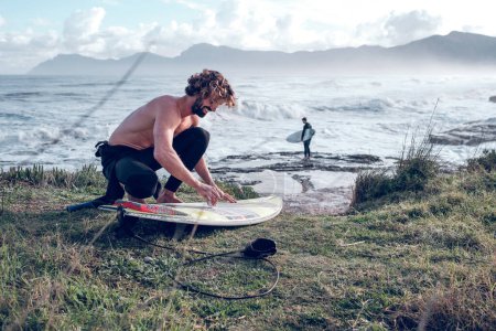 Téléchargez les photos : Vue latérale du sourire pieds nus et torse nu surfeur masculin accroupi et posant de la cire sur la planche de surf sur la côte de l'océan ondulé - en image libre de droit