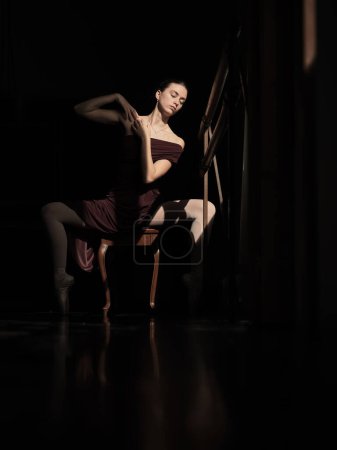 Téléchargez les photos : Corps complet de ballerine féminine en robe élégante et chaussures pointes assis en pli sur chaise dans une pièce sombre - en image libre de droit