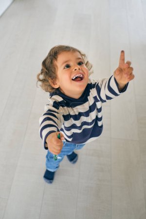 Téléchargez les photos : Corps plein angle d'adorable bambin excité avec des cheveux bouclés dans des vêtements décontractés debout sur le sol et pointant vers le haut tout en tenant marqueur coloré - en image libre de droit