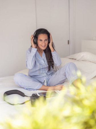 Photo for Chilling barefoot female in sleepwear sitting on cozy bed and looking away while listening to music in headphones - Royalty Free Image
