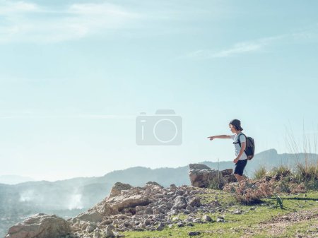 Téléchargez les photos : Vue latérale de l'enfant avec sac à dos pointant loin et admirant la crête de la montagne tout en se tenant sur la falaise contre le ciel bleu nuageux par temps ensoleillé à Majorque, Espagne - en image libre de droit