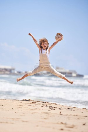 Téléchargez les photos : Corps entier de l'enfant excité dans l'ensemble sautant avec les bras levés sur le bord de mer sablonneux et tenant le chapeau à la main le jour ensoleillé d'été - en image libre de droit