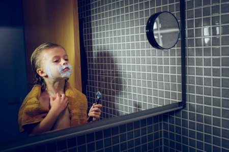 Téléchargez les photos : Garçon mignon avec serviette et mousse à raser sur le visage en regardant la réflexion dans le miroir tout en se tenant debout avec rasoir dans la salle de bain légère - en image libre de droit