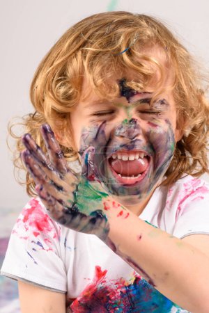 Photo for Happy dirty boy covered with colorful paints having fun while sitting in light studio with closed eyes on white background - Royalty Free Image