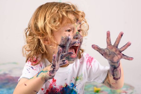 Photo for Amazed sloppy boy covered with colorful paints sitting on messy floor with opened mouth on white background in light studio - Royalty Free Image