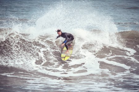 Foto de Cuerpo completo de surfista hombre en traje de neopreno de pie en la tabla de surf a caballo oleaje espumoso salpicaduras de océano en tiempo tormentoso - Imagen libre de derechos