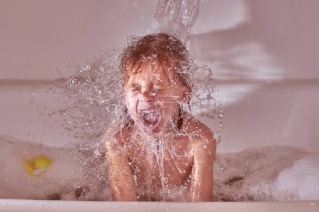 Photo for From above shirtless kid sitting in foamy water under clean stream and screaming while washing in bathroom at home - Royalty Free Image