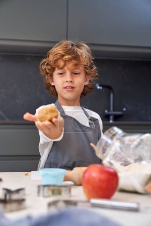 Foto de Lindo chico mirando bola de masa de galletas en la mano mientras está de pie en la mesa desordenada con varios equipos en la cocina ligera - Imagen libre de derechos