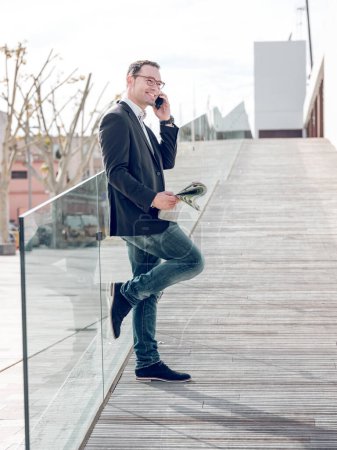Téléchargez les photos : Vue latérale de joyeux entrepreneur masculin parlant sur smartphone et discuter des questions d'affaires tout en se tenant près de clôture de verre dans la rue - en image libre de droit