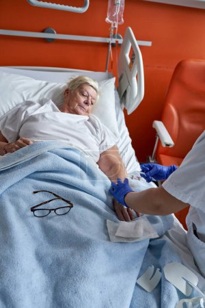 Foto de Alto ángulo de mujer anciana acostada en la cama del hospital y contemplando mientras enfermera anónima sin rostro administrando inyección intravenosa - Imagen libre de derechos