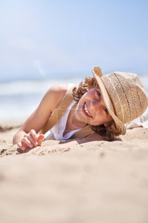 Foto de Nivel del suelo de niño preadolescente feliz en sombrero de paja tumbado en la orilla del mar y mirando a la cámara mientras disfruta de las vacaciones de verano - Imagen libre de derechos