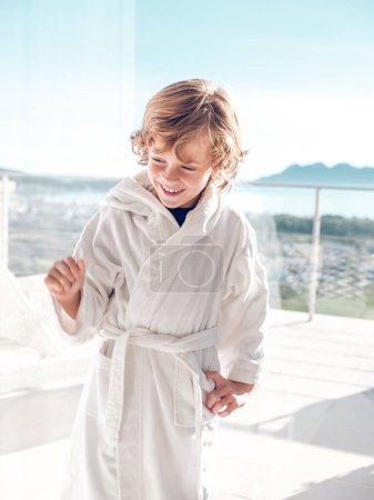 Téléchargez les photos : Joyeux adorable garçon préadolescent en peignoir éponge doux blanc profitant d'une journée ensoleillée sur la terrasse de la chambre d'hôtel dans la ville de Pollensa de la région de Majorque - en image libre de droit