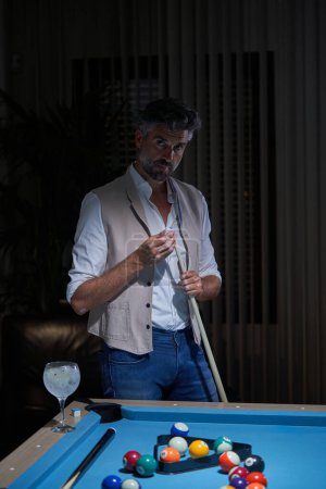 Photo for Mature billiard player looking at camera and applying chalk on cue against table with glass of cocktail in dark room - Royalty Free Image