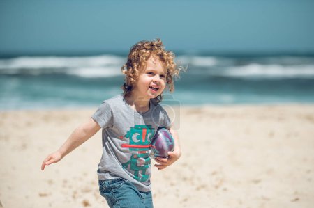 Foto de Delicioso niño preescolar en ropa casual con pelota paseando por la costa arenosa contra el mar ondulado sobre un fondo borroso en la naturaleza - Imagen libre de derechos