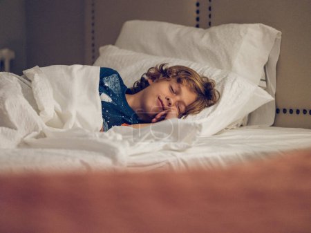 Téléchargez les photos : Enfant blond avec les cheveux bouclés touchant la joue et couché sur un lit doux tout en faisant la sieste dans la chambre d'hôtel en journée - en image libre de droit