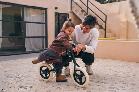 Photo for Adult dad squatting near little girl with raised leg on bicycle against house in courtyard - Royalty Free Image